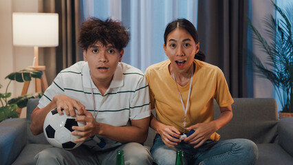 Asian new marriage couple sit on sofa excited cheering watching football sport game on television in living room at home. Young couple sport fans hold football watching online soccer on TV.