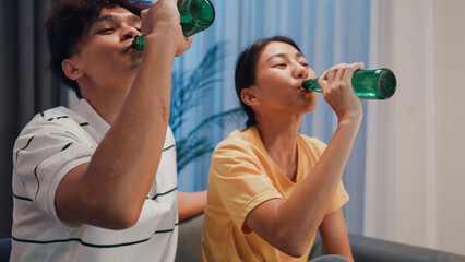 Asian new marriage couple sit on sofa excited cheering watching football sport game on television in living room at home. Young couple sport fans toast bottle beer celebration online soccer on TV.