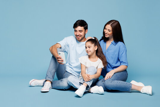 Full body young happy parents mom dad child kid daughter girl wear casual clothes sit hold in hand use mobile cell phone play together isolated on plain blue background. Family day childhood concept