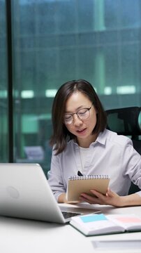 Asian businesswoman watching video call online conference taking notes using laptop at workplace in office. Female employee listening remote business training talking communicates remotely. Vertical