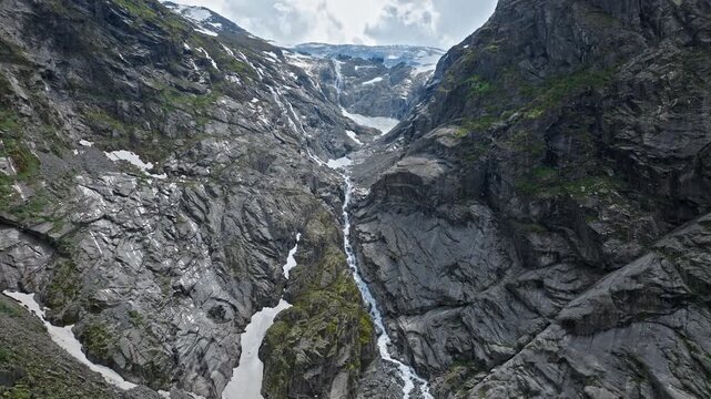 Rocky mountain gorge near Briksdalsbreen glacier in Norway. Drone view reveals steep cliffs, melting snow and flowing water in a rugged alpine environment on a summer day.