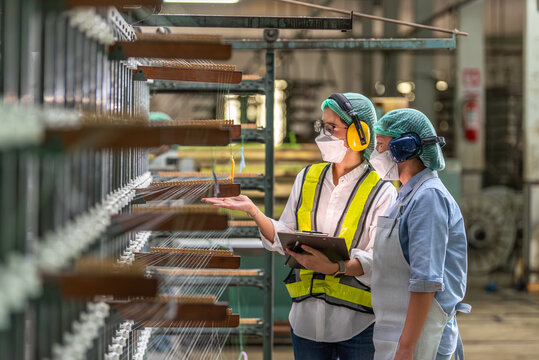 Textile inspection factory teamwork female supervisor in safety vest checking and discussing yarn production quality with woman worker in textile mill holding thread spool and clipboard.