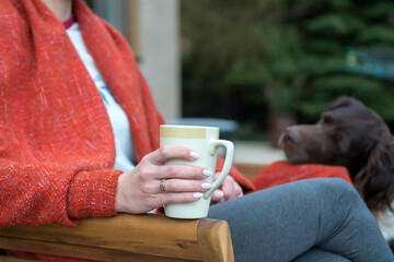 A woman holds a cup of hot drink in a cozy atmosphere. Nearby sits a dog