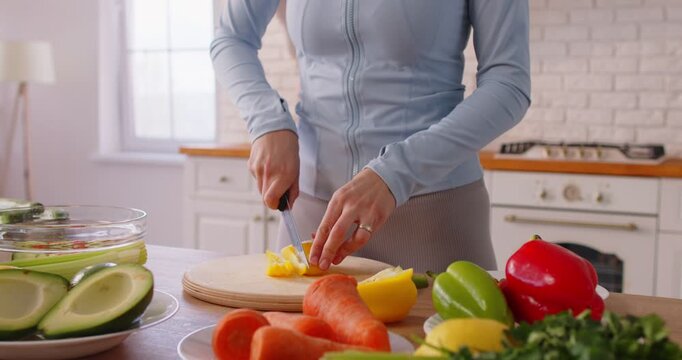 Cropped view of woman in sportswear cutting yellow bell pepper on wooden board while cooking in kitchen. Fit girl making healthy meal with organic vegetables and enjoying fresh ingredients at home.