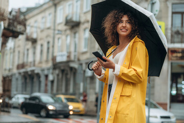 Smartphone and umbrella are in hands. Happy young woman in the city during the rain