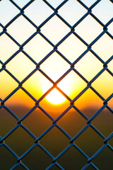Fototapeta premium Sunset seen through chain link fence, with the sun centered in diamond-shaped opening, creating dramatic warm glow and geometric pattern. Concept of separation, freedom, boundaries, security, hope