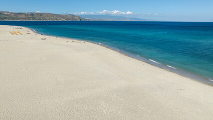 Fototapeta premium Aerial view of a vast, pristine white sand beach meeting a calm turquoise sea under a clear blue sky, with a mountain range visible in background. It is the beach of Soverato, in Calabria, Italy. 