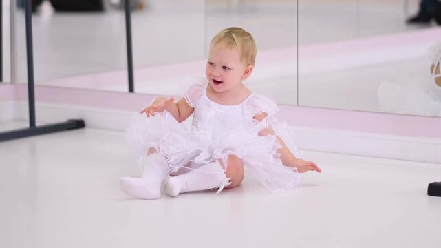 Moscow, Russia,04.04.2024 Caucasian baby girl in white tutu sitting and laughing on floor in dance studio. Childhood joy and happy moment of kid.