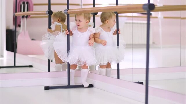 Moscow, Russia,04.04.2024 Cute baby girl in white tutu learns to walk in ballet studio with mirror and barre. Early childhood development and movement.