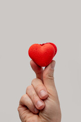 Hand gently holding a vibrant red heart-shaped macaron, symbolizing love, romance, and sweet indulgence on a clean light grey background, perfect for valentines or special occasions