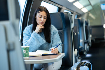 Fototapeta premium A contemplative woman takes notes in her notebook on a train while enjoying a coffee drink.