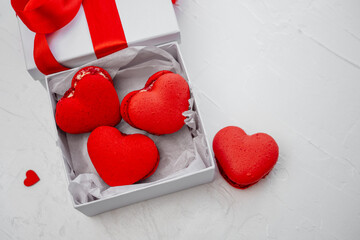 Red heart-shaped macarons resting inside a white gift box with a red ribbon, creating a sweets and dessert composition against a textured white background, symbolizing love and celebration