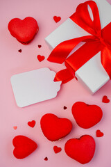 Pink background displaying a white gift box with a red ribbon and bow, surrounded by red heart-shaped macarons, confetti in heart shapes, and an empty label for a romantic message
