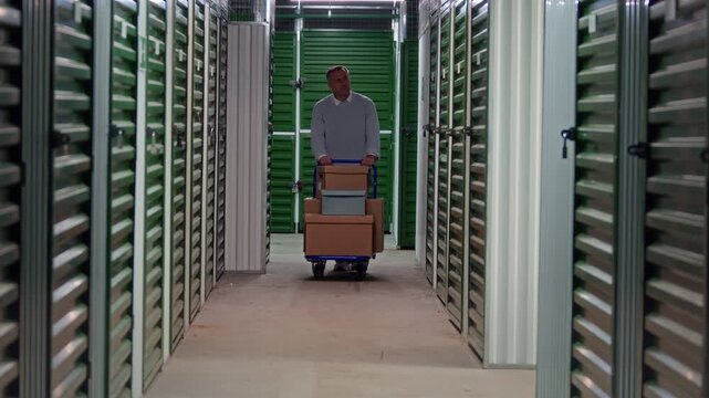 Middle aged man pushing cart loaded with cardboard boxes through corridor of secure self storage facility, searching for his unit