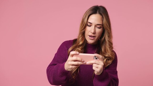 Gambling excitable young woman wear purple knitted sweater casual clothes use play racing app on mobile cell phone hold gadget smartphone for pc video games isolated on plain pastel pink background