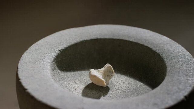 A small chalky fragment sits at the center of a rough gray stone mortar, side-lit to reveal gritty textures and a soft shadow, a quiet minimalist close-up focused on material detail.