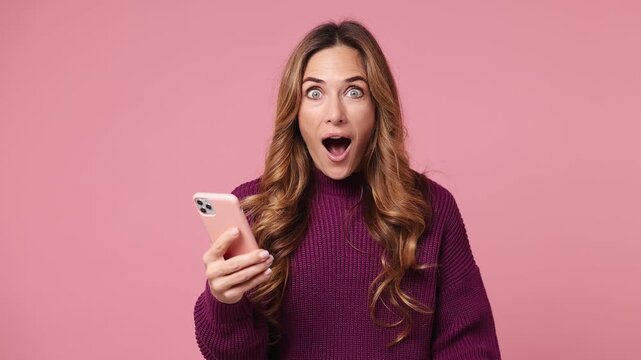 Excited happy joyful fun young woman she wear purple knitted sweater casual clothes use mobile cell phone hold win fan of cash money in dollar banknotes isolated on plain pastel light pink background