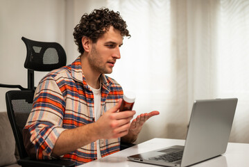Young man showing pill bottle to doctor on his laptop, asking about the correct dose to manage his medical condition while sitting at home. Telemedicine concept.	