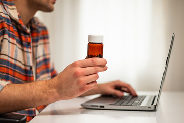 Close up photo of man holding pill bottle and using laptop for searching more information about his prescription medicine while sitting at his home. Focus on pill bottle.	