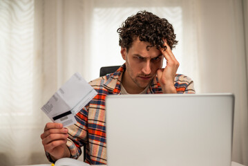 Stressed man using laptop and credit card to pay his financial bills online while sitting at home. He is having worried expression on face while managing his budget and expenses.