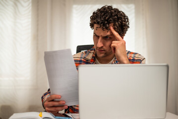 Stressed businessman looking and analyzing documents while working on laptop at his home office.	