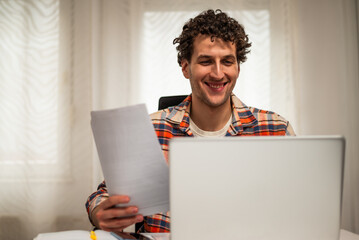 Cheerful young freelancer using laptop and checking documents while working at his cozy home.	