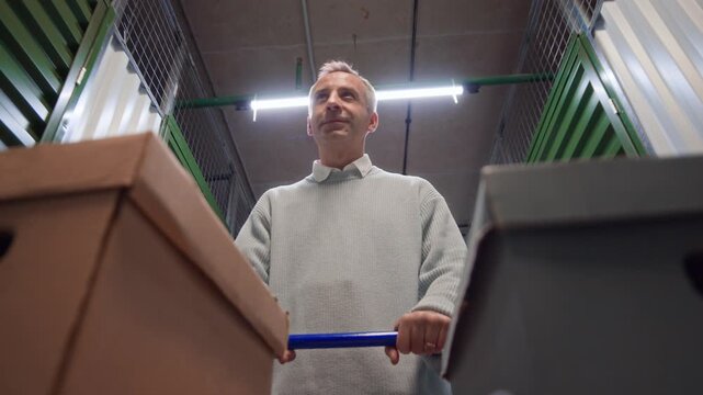 Low angle view of middle aged man pushing cart full of cardboard boxes while walking through self storage hallway