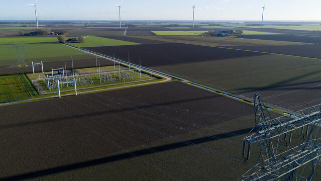 Aerial view of power lines casting long shadows across rich, dark earth, meeting the horizon where wind turbines stand tall, Dronten, Flevoland, Netherlands.