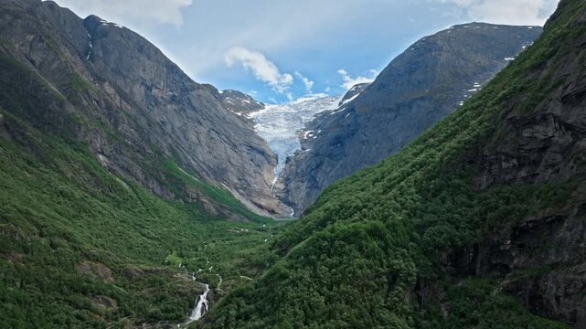 Glacier valley at Briksdalsbreen in Norway surrounded by mountains. Drone view captures green hillsides, rocky cliffs and melting ice under bright summer light.