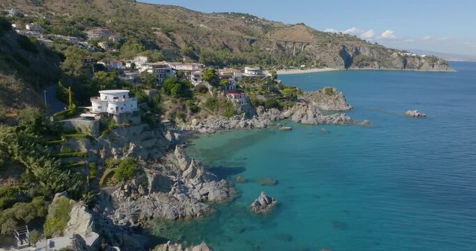 High-angle aerial view of a rugged coastline where colorful houses are built into the rocky cliffs, overlooking clear turquoise waters and a secluded beach nestled at the foot of a verdant hillside.
