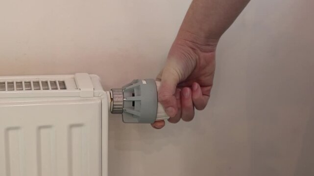 A woman's hand turns a mechanical thermostat on a radiator. A close-up of a temperature regulator on a battery. A mechanical thermal head of a battery regulator with a scale from 0 to 5