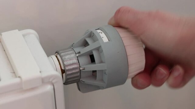 A woman's hand turns a mechanical thermostat on a radiator. A close-up of a temperature regulator on a battery. A mechanical thermal head of a battery regulator with a scale from 0 to 5