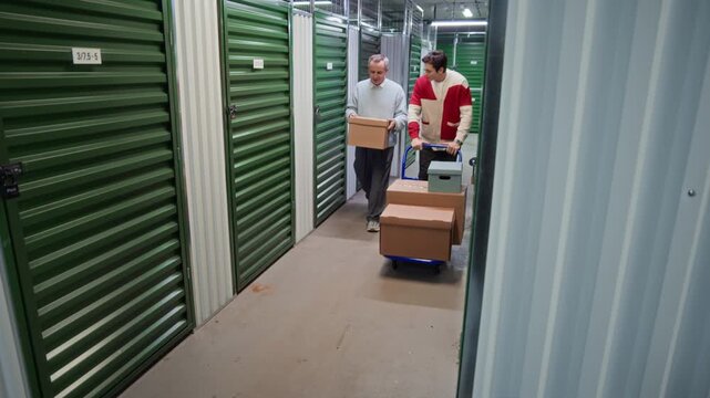 Slow motion shot of middle aged man carrying cardboard box and talking to his son pushing loaded cart down corridor while walking together through storage facility