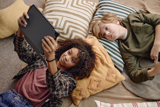 Two Generation Alpha kids lying on pillows using digital tablet together, Black girl holding device while both looking at screen, casual indoor setting