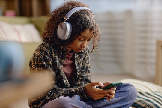 Black teenager girl sitting on floor using smartphone while wearing large headphones, focusing on screen, engaging with digital content, representing Generation Alpha with gadget
