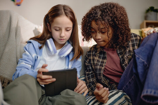 Two Generation Alpha kids sitting together using digital tablet, Caucasian girl with headphones and Black girl focusing on screen, engaging with modern gadget technology indoors