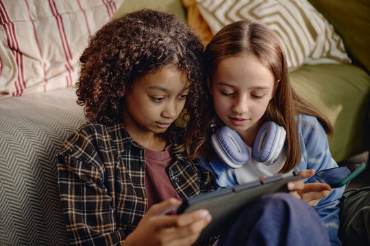 Two Generation Alpha girls sitting closely together using digital tablet, focusing on screen, sharing headphones, engaging with modern gadget technology