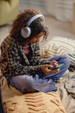 Black preteen girl sitting cross legged using smartphone while wearing wireless headphones, focusing on screen, engaging with digital content, surrounded by cushions indoors