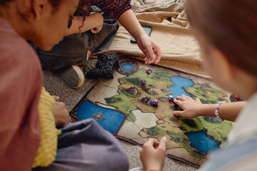 Multiethnic group of Generation Alpha kids sitting on floor playing tabletop board game with fantasy map and dice, hands moving game pieces, engaging in strategy © Seventyfour