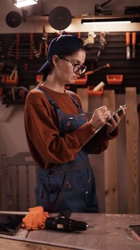 Side view of female handyman in blue overalls writing on clipboard in small garage or home workshop