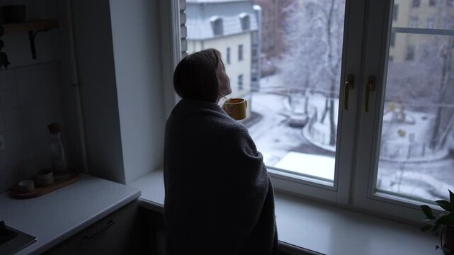Woman wrapped in blanket standing by window with mug, watching snowy city street, calm reflective winter morning. Peaceful female spending mindful time view from window, holding warm drink in hands.