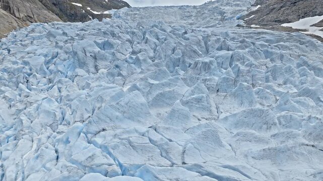 Aerial view flying over Briksdalsbreen glacier in Norway. Vast blue ice and rugged formations stretch across the frame, emphasizing dramatic alpine wilderness and climate scenery.