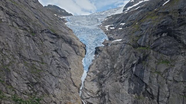 Glacier tongue cutting through steep rock walls at Briksdalsbreen in Norway. Aerial drone view highlights dramatic ice flow, cliffs and alpine terrain in summer.