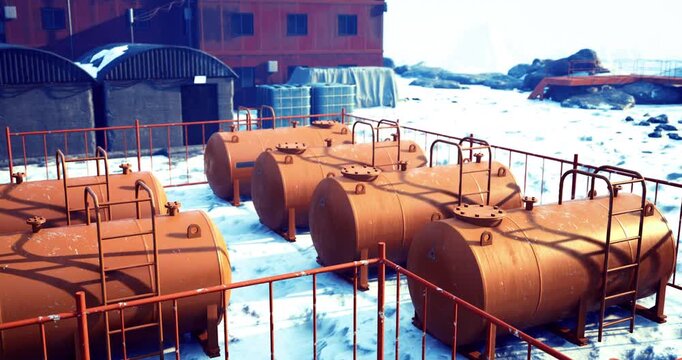 Storage tanks are arranged in a row on a snowy ground next to a red building in a frigid area. The scene includes industrial structures and a clear blue sky.