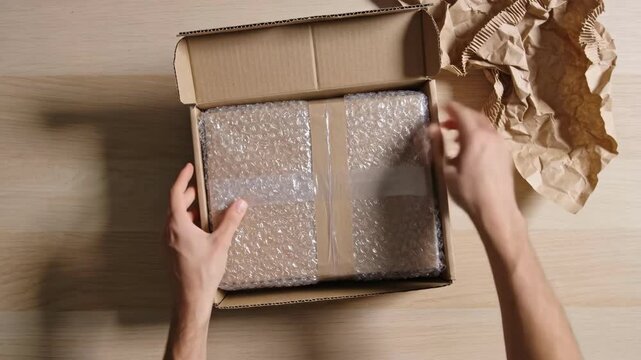 Eco friendly packaging scene shows hands using kraft paper cushioning as void fill around bubble wrapped product, preparing parcel for delivery with reduced plastic waste concept.