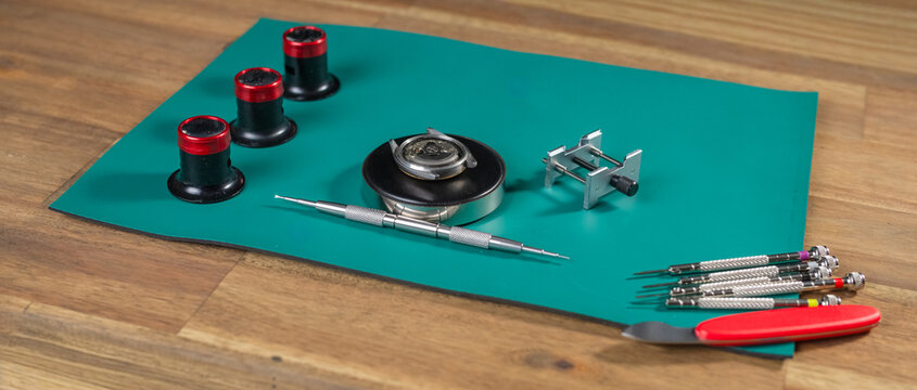Tools and equipment for watch repair set on a green mat indicate the work of a skilled watchmaker focusing on fine mechanics.