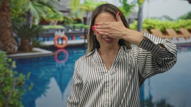 Woman wearing striped shirt covers eyes with hand by pool building; shyness introspection solitude vulnerability.