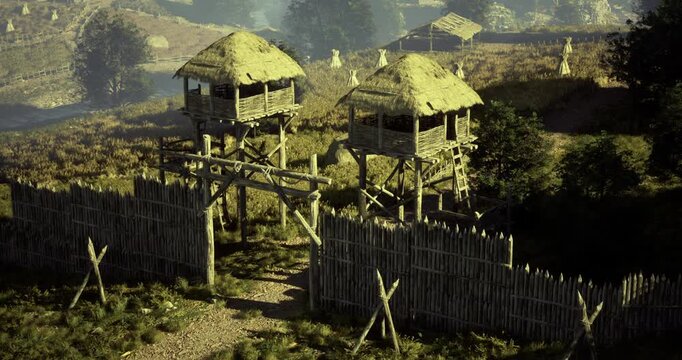 Two wooden watchtowers stand guard over an ancient settlement surrounded by lush greenery and clear skies. A dirt path leads through a wooden gate, showcasing a peaceful environment.