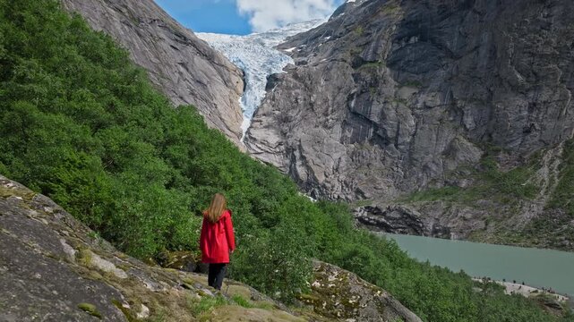 Young woman in red jacket standing near Briksdalsbreen valley in Norway. She looks toward the glacier and mountains on a sunny summer day, enjoying nature and adventure travel.