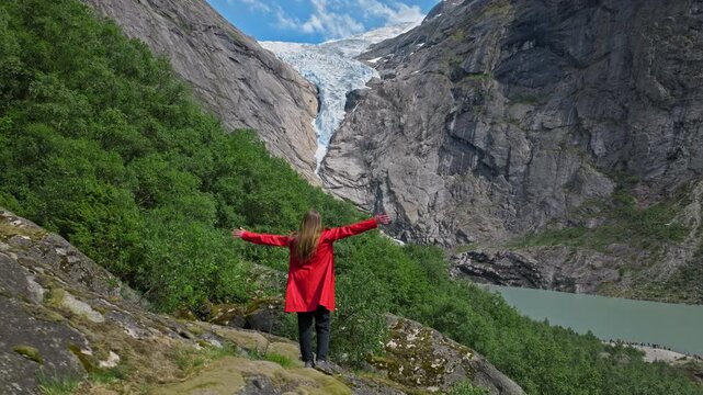 Woman in red jacket raising arms toward Briksdalsbreen glacier in Norway. She celebrates the dramatic mountain scenery and outdoor adventure on a bright summer day.